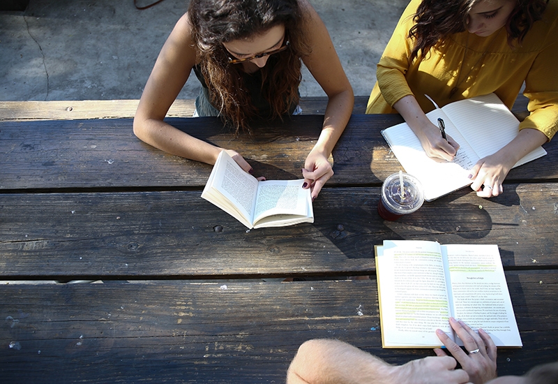 Three adults sit at outdoor table, reading books and taking notes
