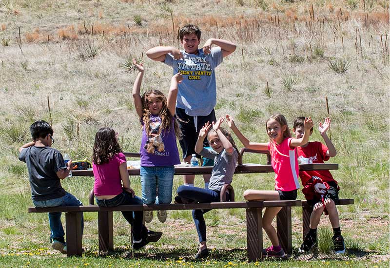 Students sit, smiling, at outdoor picnic table with arms raised in the air
