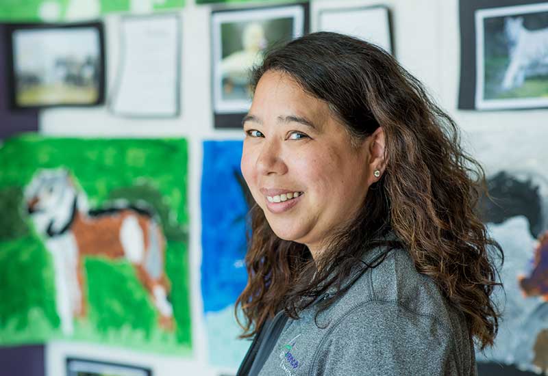 Teacher smiles at camera with student artwork on display in background
