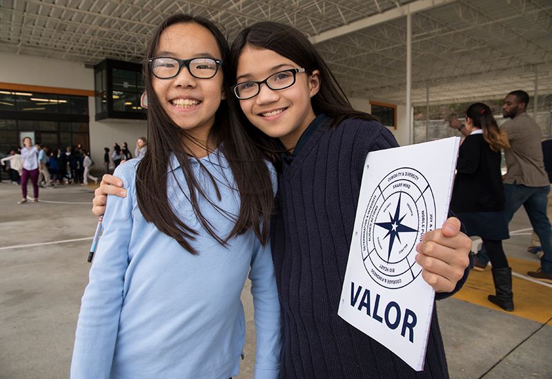 Two students stand close together, one with arm draped over the other's arm, holding Valor school sign
