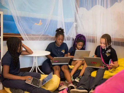 Four female students sit on floor, bean bag chairs while working on laptops
