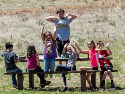 Students sit, smiling, at outdoor picnic table with arms raised in the air
