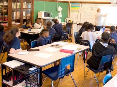 Teacher stands in front of white board inside classroom while students in seats look on

