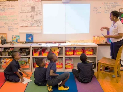 Four students sit on colorful floor, looking up at teacher as she gestures at white board
