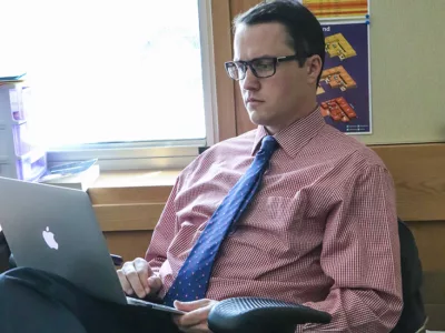 Teacher sits in chair in classroom, alone, looking down at laptop
