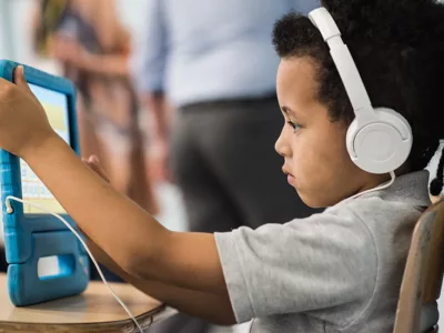 Elementary student sits at desk with headphones on while holding tablet in front of him
