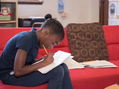 Student sits on red couch, leaning forward as she writes on paper
