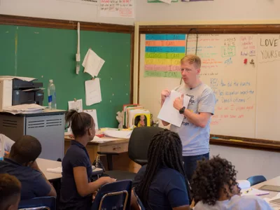 Teacher stands in front of classroom, pointing to paper in his hand as students look on
