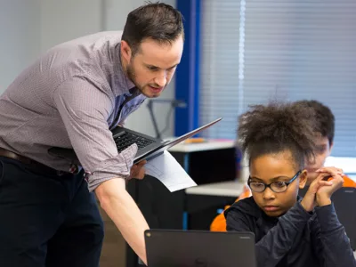 Teacher, holding laptop, works with student as she works on her own laptop
