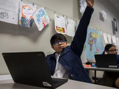 Student raises hand, sitting at desk
