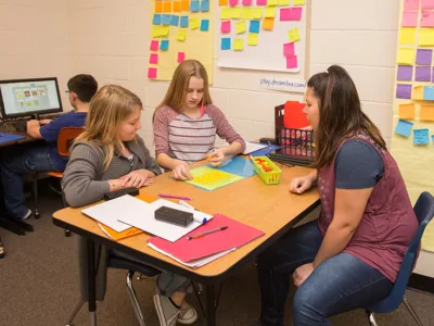 Teacher works with pair of students at small desk
