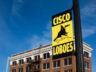 Outdoor view of school building, with a yellow and black sign reading "Cisco Loboes" in the foreground, showing the school's mascot
