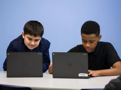 Two students work side-by-side at table on laptops, smiling
