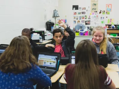 Students sit together in classroom, looking at laptops, while one looks up and raises his hand
