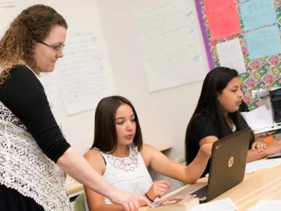 Photo of teacher standing and speaking with seated student working on her laptop
