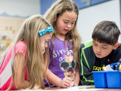 Three elementary students working together at table with several resources
