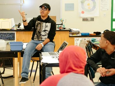 Student speaks, gesturing, while two students look on
