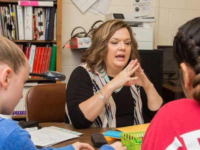 Teacher sits at desk, gesturing as she speaks
