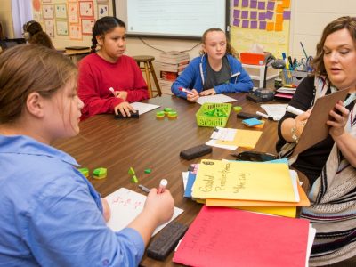 Teacher holds up clipboard, showing it to students at table as they look on carefully

