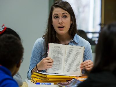 Teacher holds open book, presenting it to students, talking
