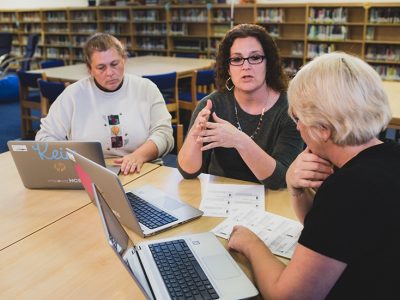 Three teachers sit beside each other in library, talking, with papers and laptops around them
