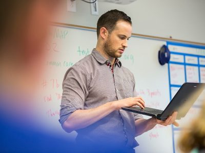 Teacher holds laptop in hand, typing, standing in classroom
