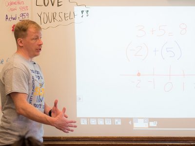 Teacher stands in front of whiteboard, looking out at class
