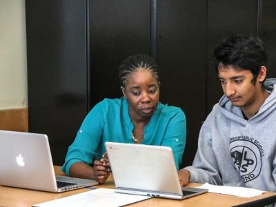 Photo of teacher and student working side by side at table with laptops directly in front of them
