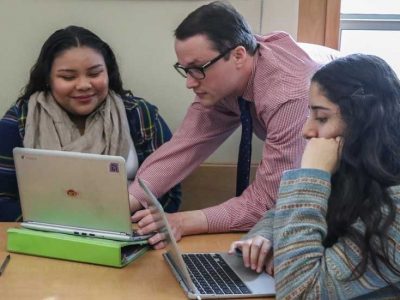 Photo of teacher working with two students on their laptops
