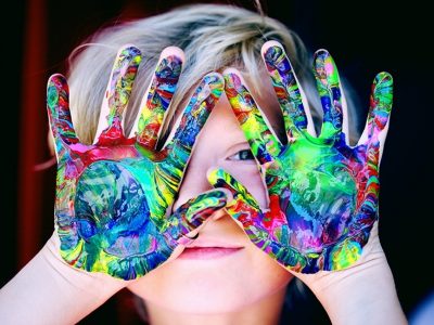A child's raised hands with bright, multicolored swirls of paint on both palms with the child's eyes peeking through their spread fingers.
