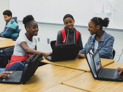 Three students, smiling, work together off of one laptop
