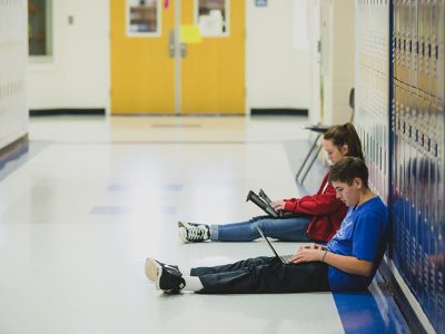 Two students lean against lockers while working off of laptops
