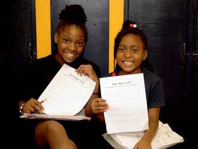 Two students sit beside each other in hallway and hold up reports, smiling
