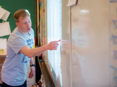 Teacher stands in front of interactive white board, pointing at the screen
