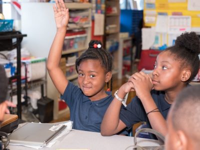 Two female students sit at desk in classroom as one raises her hand
