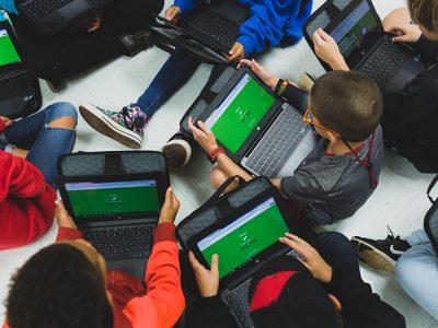 Students sit on floor with laptops in laps, screens are showing the same green background
