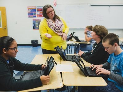 Teacher speaks with student while he sits at joined desks with other students who are looking at their laptops
