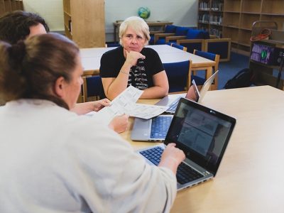 Three teachers sit at desk with laptops, holding papers, speaking with each other
