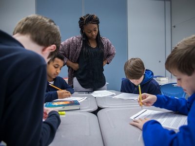 Teacher watches as students work at shared desks on paper
