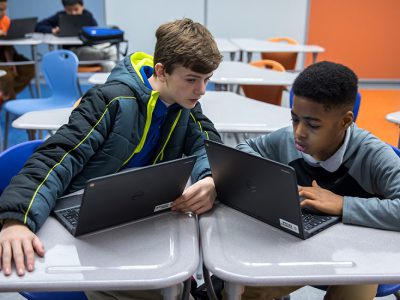 Two students in classroom using laptops, while looking at one student's screen
