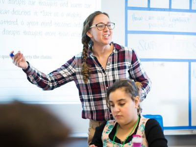 Teacher stands in front of white board, behind student, and smiles as she gestures to classroom
