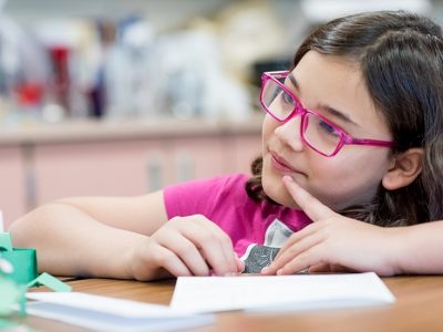 Girl wearing glasses looks off to the side, in a thinking pose, holding a pencil to a paper on a desk

