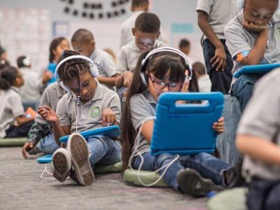 Photo of multiple students seated at desks and on floor using devices with headphones
