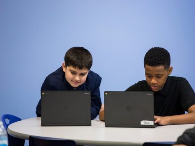 Two students in front of blue wall, sitting at desk, looking at laptops
