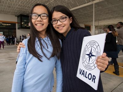 Two students stand close together, one with arm draped over the other's arm, holding Valor school sign
