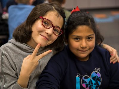 Two students, one showing a peace sign, draping her arm around another student's shoulders
