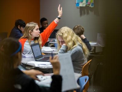 Student raises her hand, sitting among classmates
