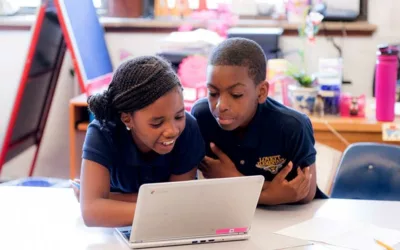 Two students work inside classroom, looking at laptop together