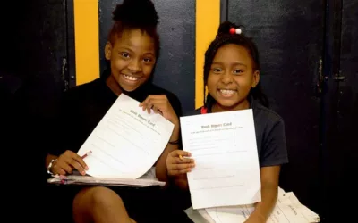 Two students sit beside each other in hallway and hold up reports, smiling