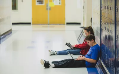 Two students lean against lockers while working off of laptops